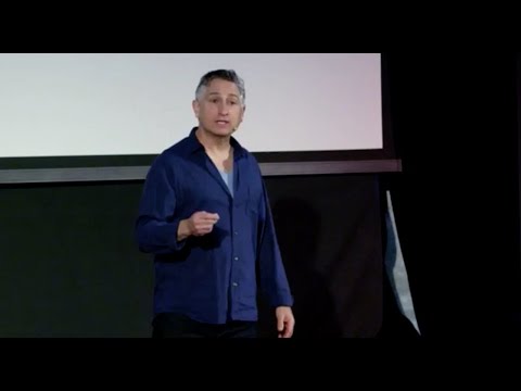 Ein Mann in einem blauen Hemd mit Knöpfen, der auf der Bühne vor einer Leinwand gestikuliert, während er spricht.