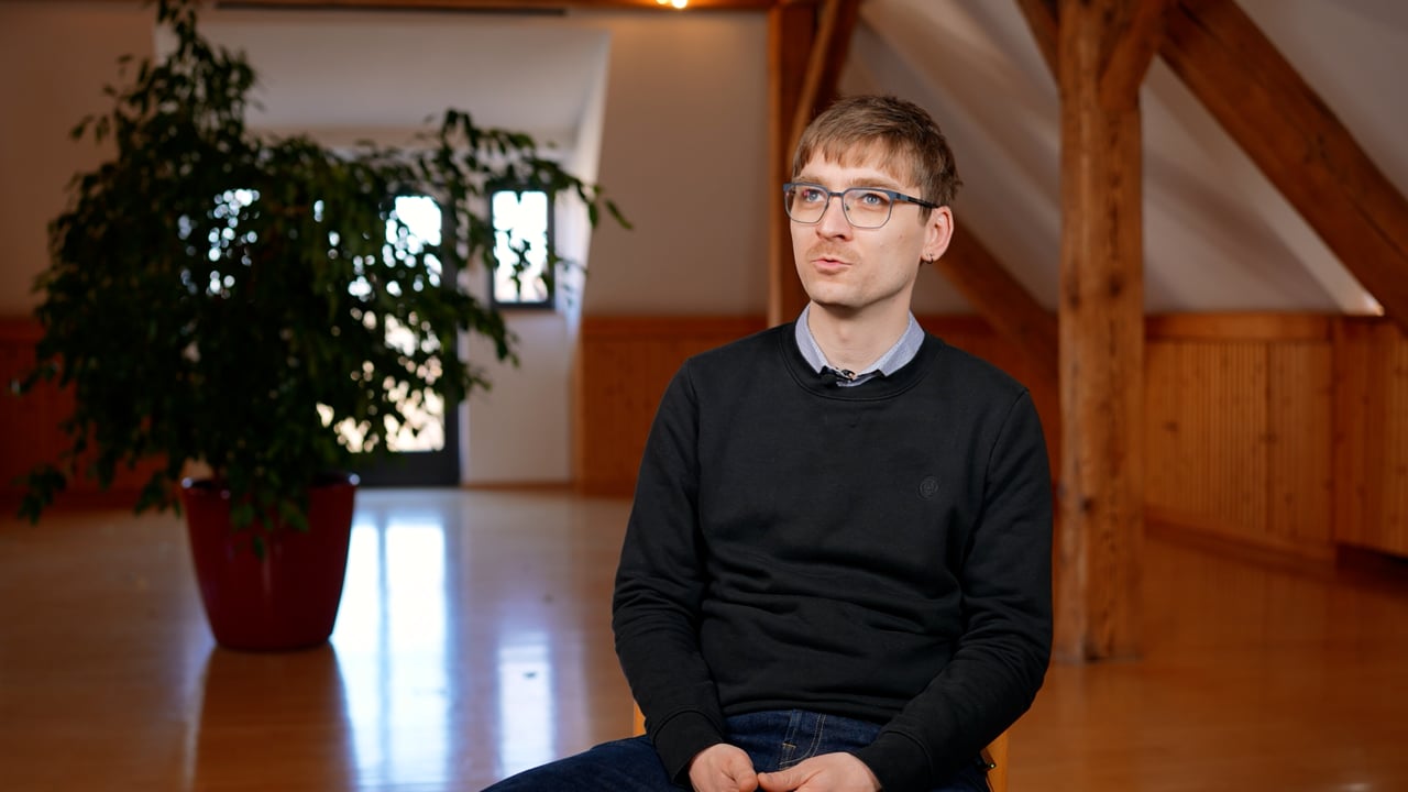 Eine Person mit Brille und einem dunklen Pullover sitzt drinnen auf einem Holzboden; im Hintergrund sind eine große Pflanze und Holzbalken zu sehen.