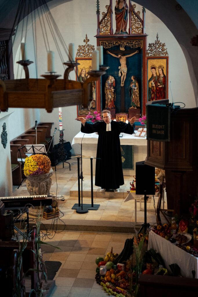 Ein Priester in schwarzer Robe steht mit erhobenen Armen vor einem Altar in einer reich geschmückten Kirche, umgeben von Blumen, Obst und Gemüse.