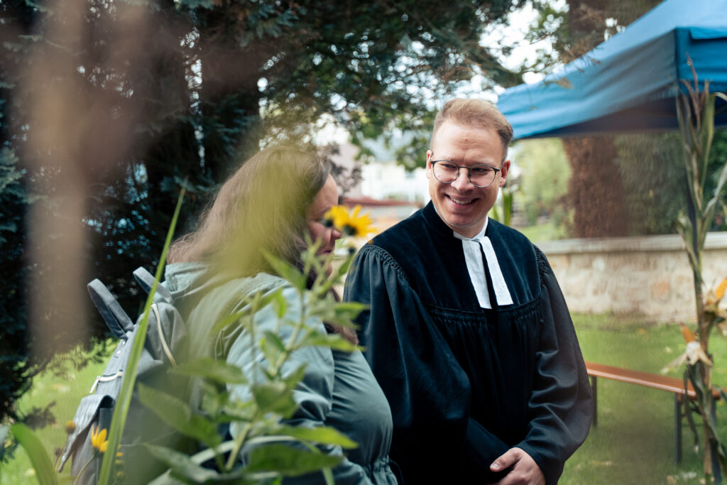 Ein lächelnder Mann in einer schwarzen Robe mit weißem Kragen steht im Freien und unterhält sich mit einer Frau mit braunen Haaren und einer grünen Jacke. Im Hintergrund sind Bäume und ein blaues Vordach zu sehen.