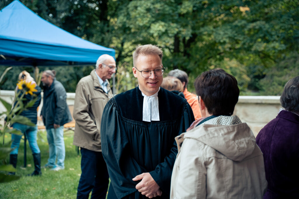 Ein Mann in einer schwarzen Robe und einem weißen Kragen lächelt und unterhält sich mit einer Frau im Freien; mehrere Menschen stehen in der Nähe unter einem blauen Vordach, im Hintergrund sind Bäume zu sehen.
