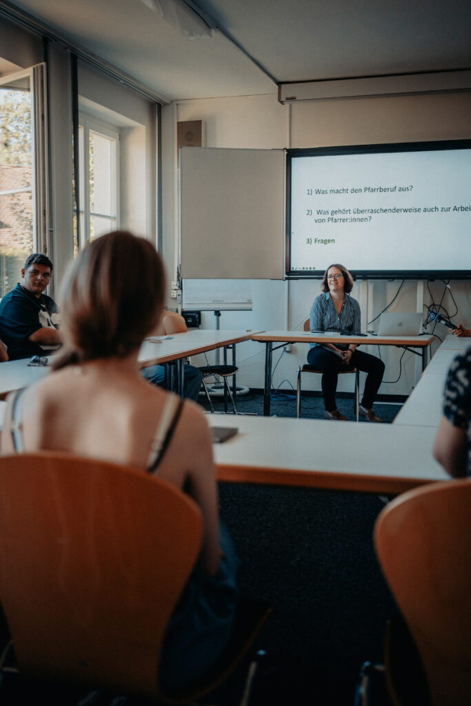 Eine Gruppe von Menschen sitzt in einem Klassenzimmer und blickt zu einer Frau, die eine Diskussion leitet. Auf einer Leinwand hinter ihr sind drei Fragen auf Deutsch zu sehen.