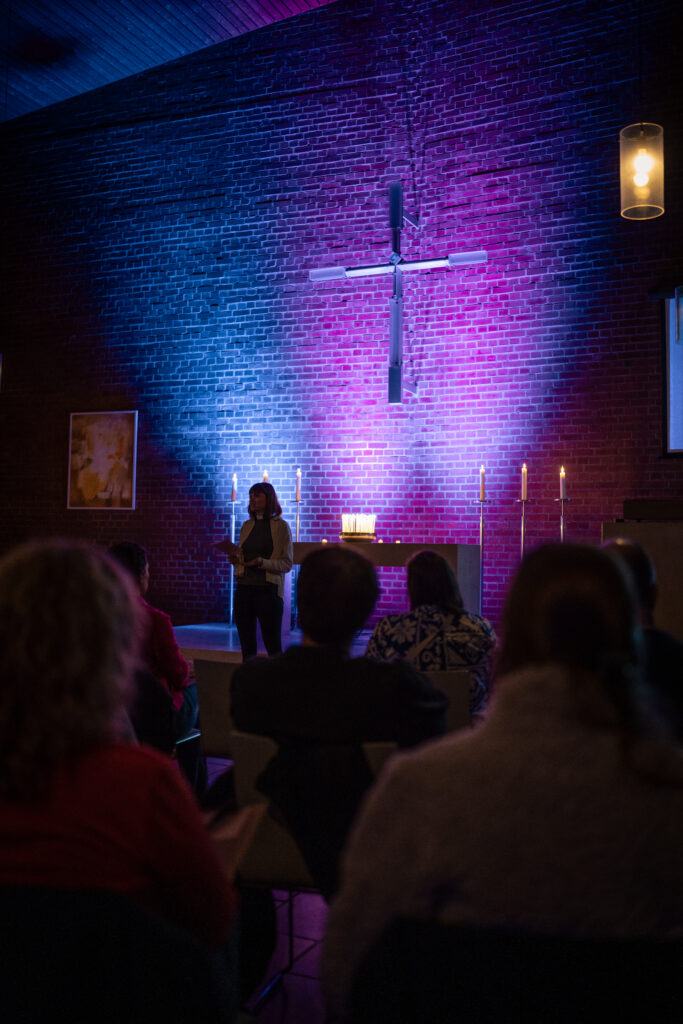 Eine Person spricht vor einem sitzenden Publikum in einer schummrigen Kapelle, in der ein Kreuz, Kerzen und violett-blaue Beleuchtung an einer Backsteinwand zu sehen sind.