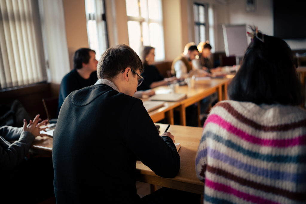 Die Menschen sitzen in einem sonnendurchfluteten Klassenzimmer an Tischen, schreiben und hören zu, während auf den Tischen Hefte und Blätter liegen.