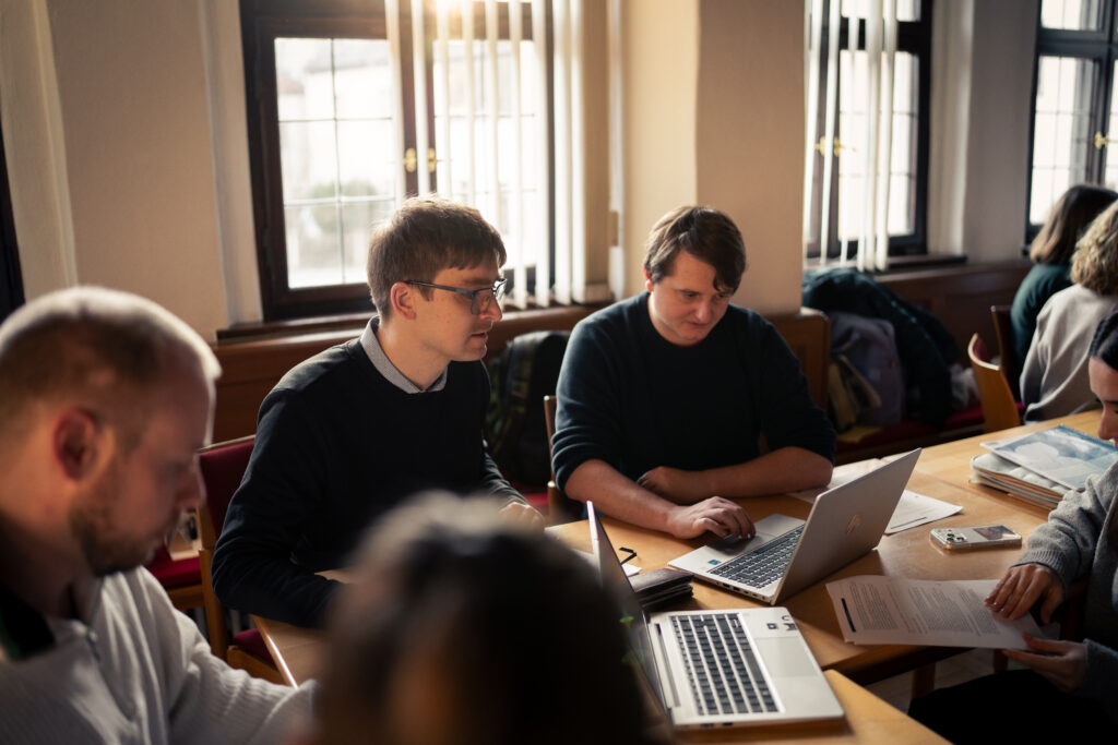 Vier Personen arbeiten gemeinsam an einem Tisch mit Laptops und Unterlagen in einem sonnendurchfluteten Raum mit großen Fenstern und Vertikaljalousien.