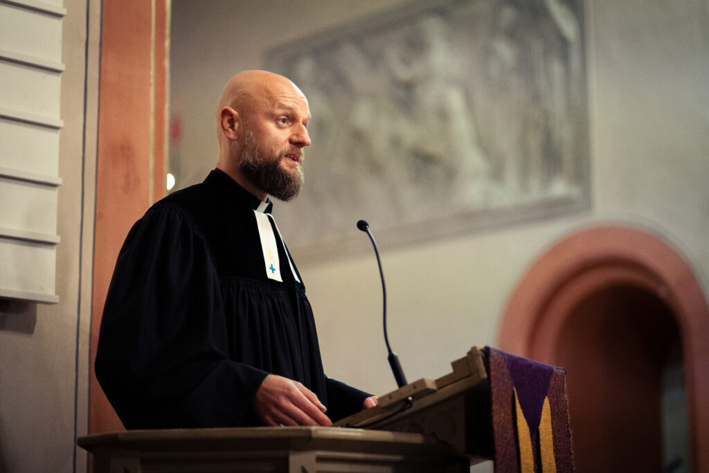Ein Mann in einer schwarzen Robe hält in einer Kirche eine Rede am Rednerpult, das mit einem Mikrofon und einem violetten Tuch drapiert ist. Im Hintergrund ist ein Wandgemälde zu sehen.