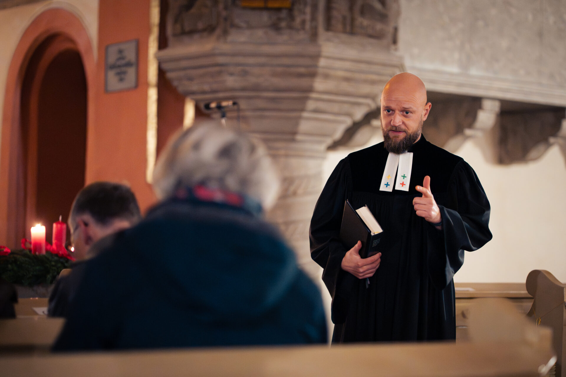 Ein Pfarrer in einer schwarzen Robe hält ein Buch in der Hand und gestikuliert, während er zu den Menschen spricht, die in den Kirchenbänken sitzen.