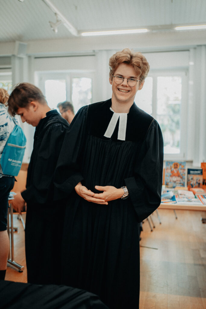 Ein junger Mensch in einer schwarzen Robe mit weißem Kragen lächelt in einem hellen Raum, im Hintergrund sind Bücher und Menschen zu sehen.