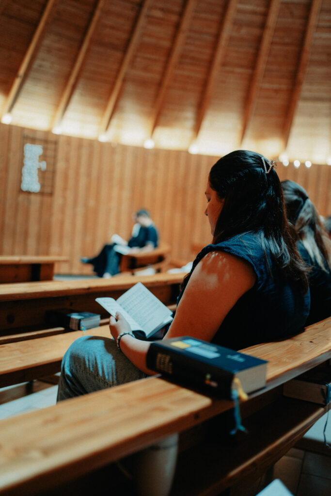 Eine Frau sitzt auf einer Holzbank in einer Kirche und liest ein Buch, während im Hintergrund eine weitere Person liest. Licht fällt durch die Holzdecke.