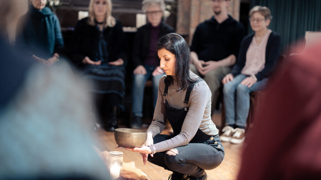 Eine Frau kniet auf dem Boden und hält eine Klangschale in der Hand; um sie herum sitzen Menschen in einem sanft beleuchteten Raum, was auf eine Gruppenmeditation oder eine Achtsamkeitssitzung hindeutet.