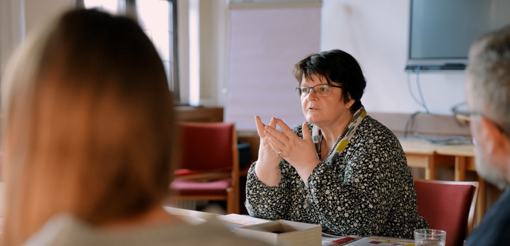Eine Frau mit Brille sitzt an einem Tisch und spricht, während sie mit den Händen gestikuliert, zu zwei Personen in einem Besprechungsraum, in dem Stühle und ein Flipchart stehen.
