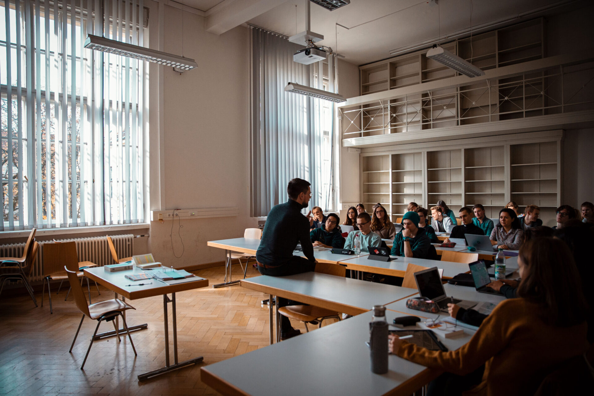 Ein Lehrer sitzt auf einem Pult vorne im Klassenzimmer und spricht zu den Schülern, die sitzen und mit ihren Laptops und Notizbüchern aufmerksam zuhören.