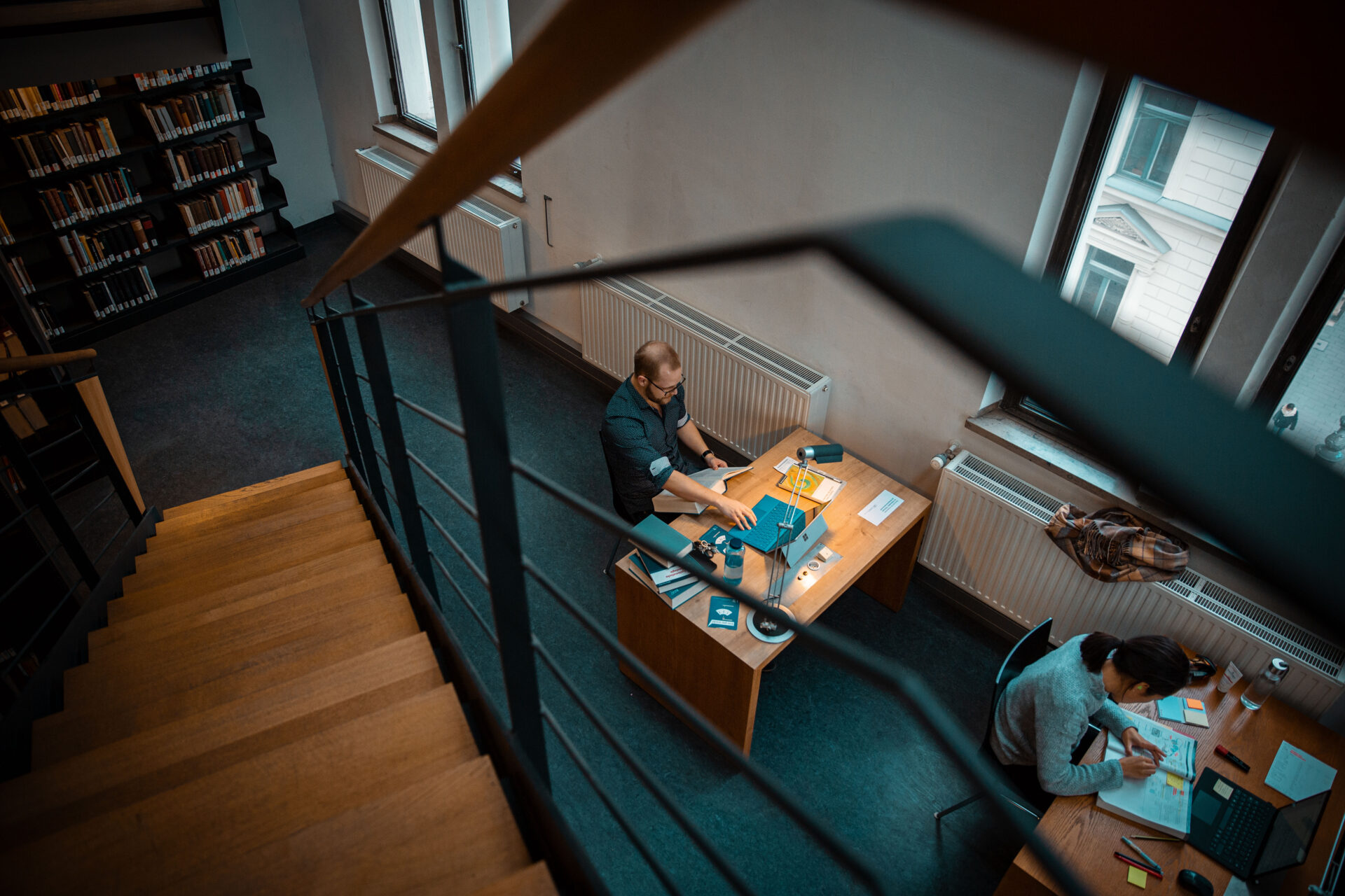 Zwei Personen lernen an getrennten Schreibtischen in einer Bibliothek neben den Fenstern. Die Aufnahme wurde von oben in der Nähe einer Treppe gemacht; im Hintergrund sind Bücherregale zu sehen.