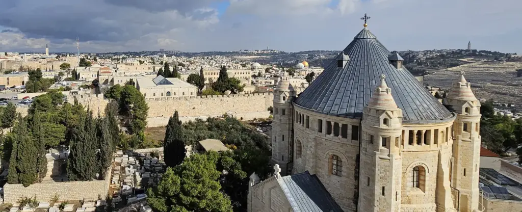Ein Panoramablick auf Jerusalem mit historischen Steingebäuden, einer Kuppelkirche, Stadtmauern, Bäumen und einem Friedhof unter einem teilweise bewölkten Himmel.