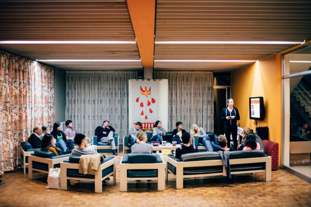 Eine Gruppe von Menschen sitzt im Halbkreis in einem Aufenthaltsraum, während eine Person steht und mithilfe einer Leinwand vorne im Raum eine Präsentation hält.