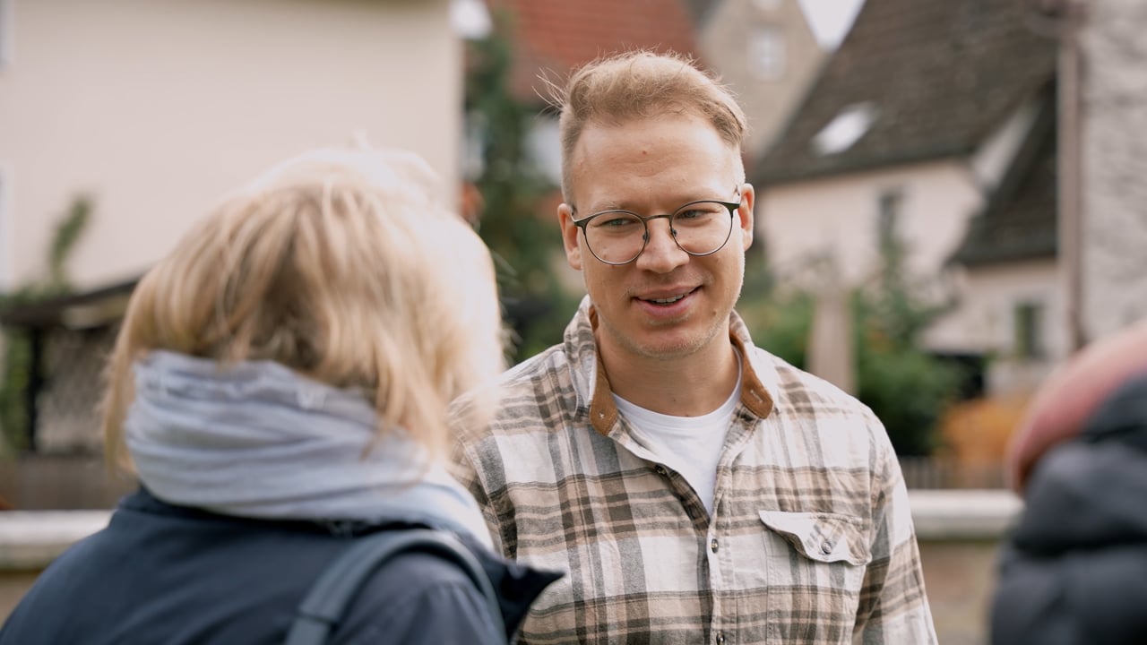 Ein Mann mit Brille und kariertem Hemd unterhält sich draußen in einem Wohngebiet mit einer Frau mit blonden Haaren und einem blauen Schal.