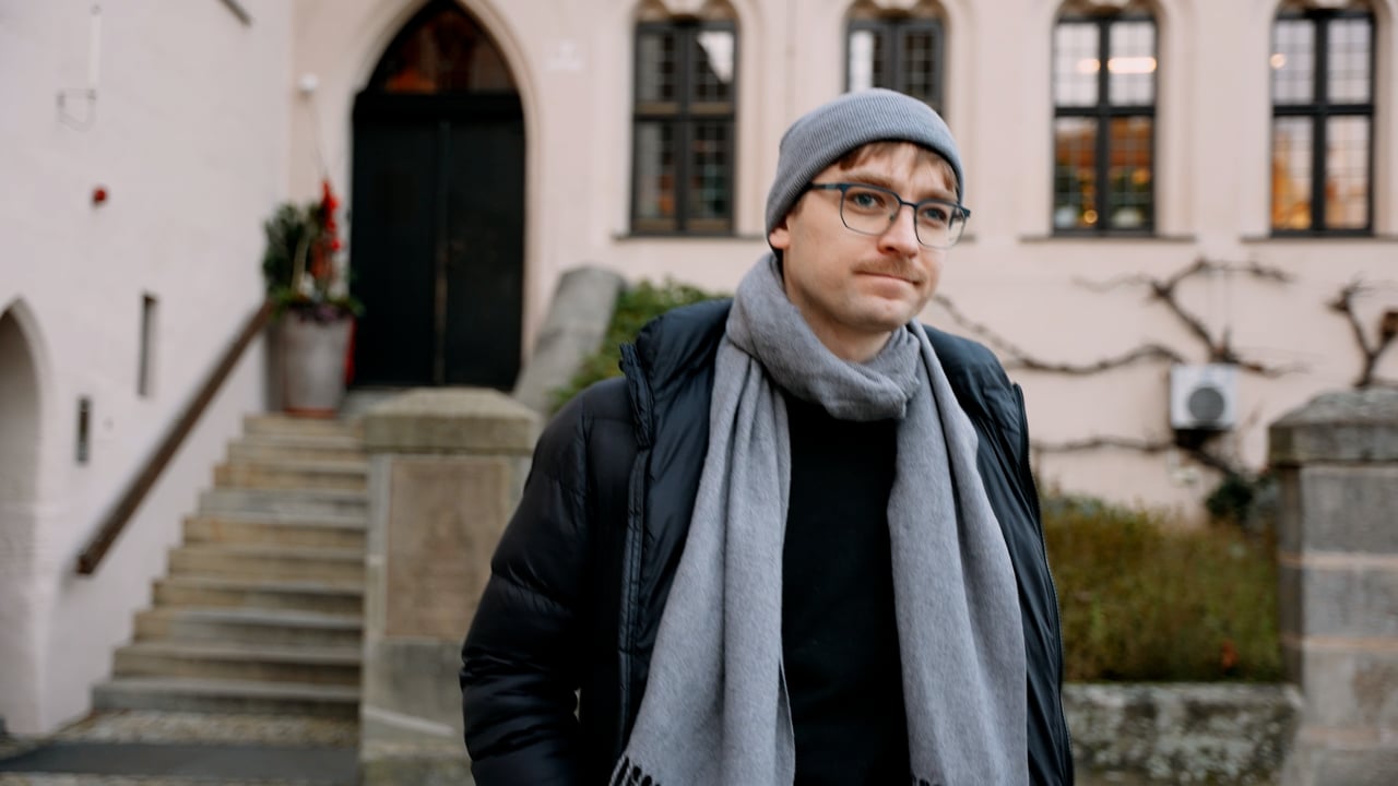 Ein Mann mit Brille, grauer Mütze und Schal steht vor einem Gebäude mit Bogenfenstern und einer Treppe und blickt leicht zur Seite.