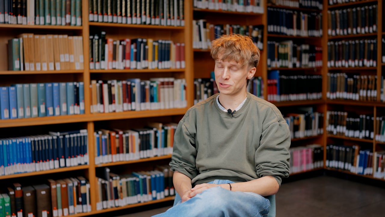 Eine Person mit hellem Haar und einem grünen Sweatshirt sitzt in einer Bibliothek vor einem Bücherregal voller Bücher und blickt mit geschlossenen Augen nach unten.