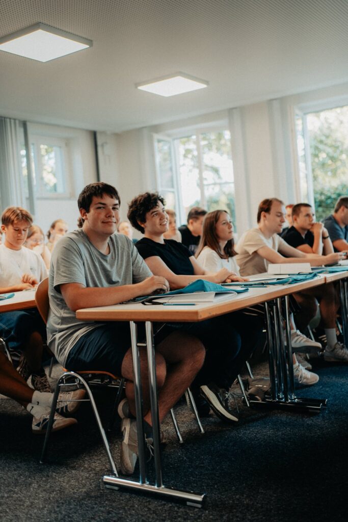 Die Schüler sitzen an ihren Schreibtischen in einem Klassenzimmer, hören aufmerksam zu und machen sich Notizen, während im Hintergrund Tageslicht durch die großen Fenster hereinströmt.