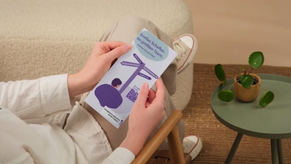 Eine Person sitzt auf einem Sofa und hält eine Broschüre mit abgebildeten Wegweisern und Text in der Hand. Auf einem runden Tisch in der Nähe steht eine kleine grüne Pflanze.