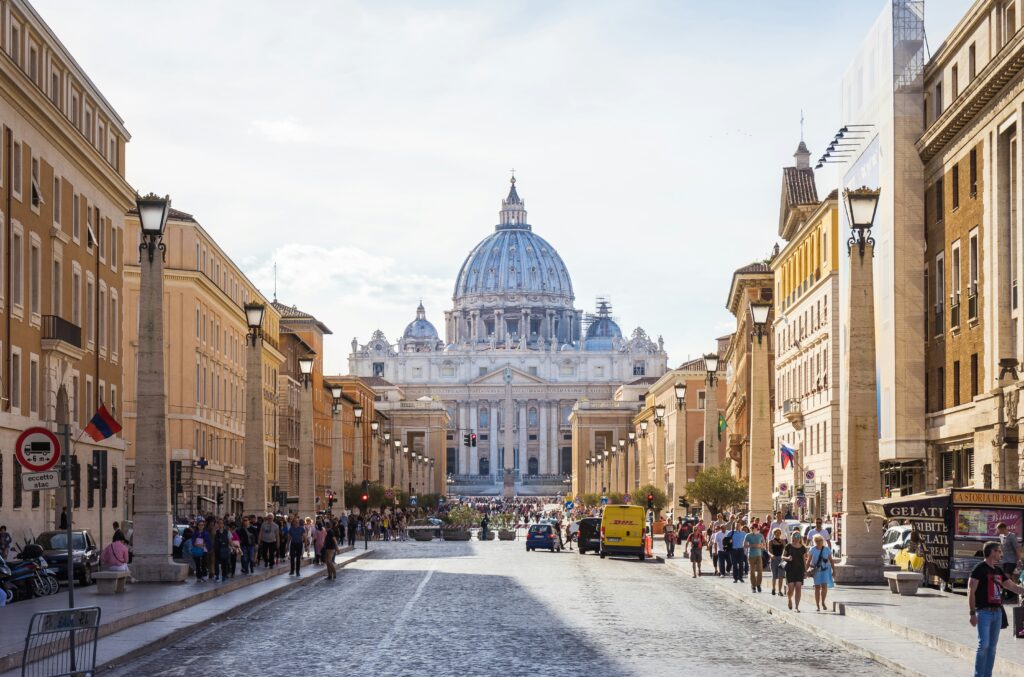 Unter strahlend blauem Himmel schreitet eine Menschenmenge eine von Gebäuden gesäumte Kopfsteinpflasterstraße entlang in Richtung des Petersdoms im Vatikan.