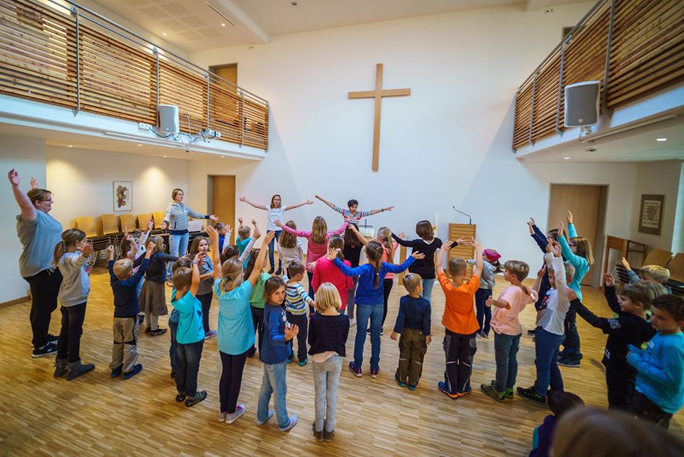 Die Kinder stehen im Kreis, während Erwachsene in einem hellen Raum mit einem großen Kreuz an der Wand und Holzböden eine Aktivität leiten.
