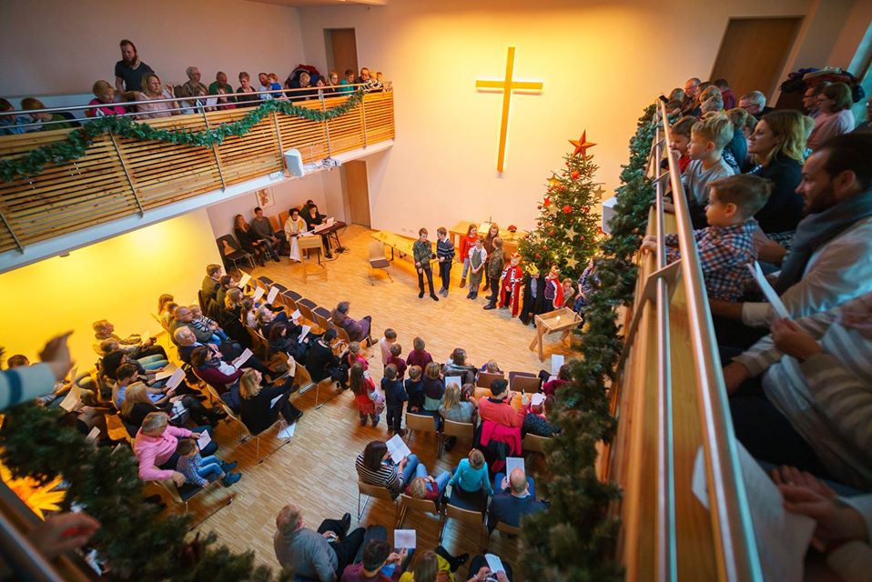 Die Menschen versammeln sich in einer mit Girlanden und einem Weihnachtsbaum geschmückten Kirche und sehen zu, wie Kinder in der Nähe eines großen Kreuzes singen. Einige sitzen unten, andere schauen von einem Balkon aus zu.