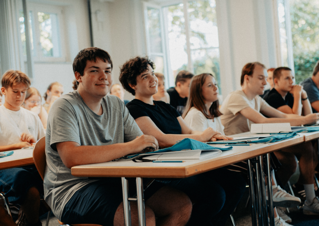Die Schüler sitzen an ihren Schreibtischen in einem hellen Klassenzimmer; einige lächeln in die Kamera, vor sich haben sie Bücher und Schultaschen, und durch die Fenster fällt Tageslicht herein.