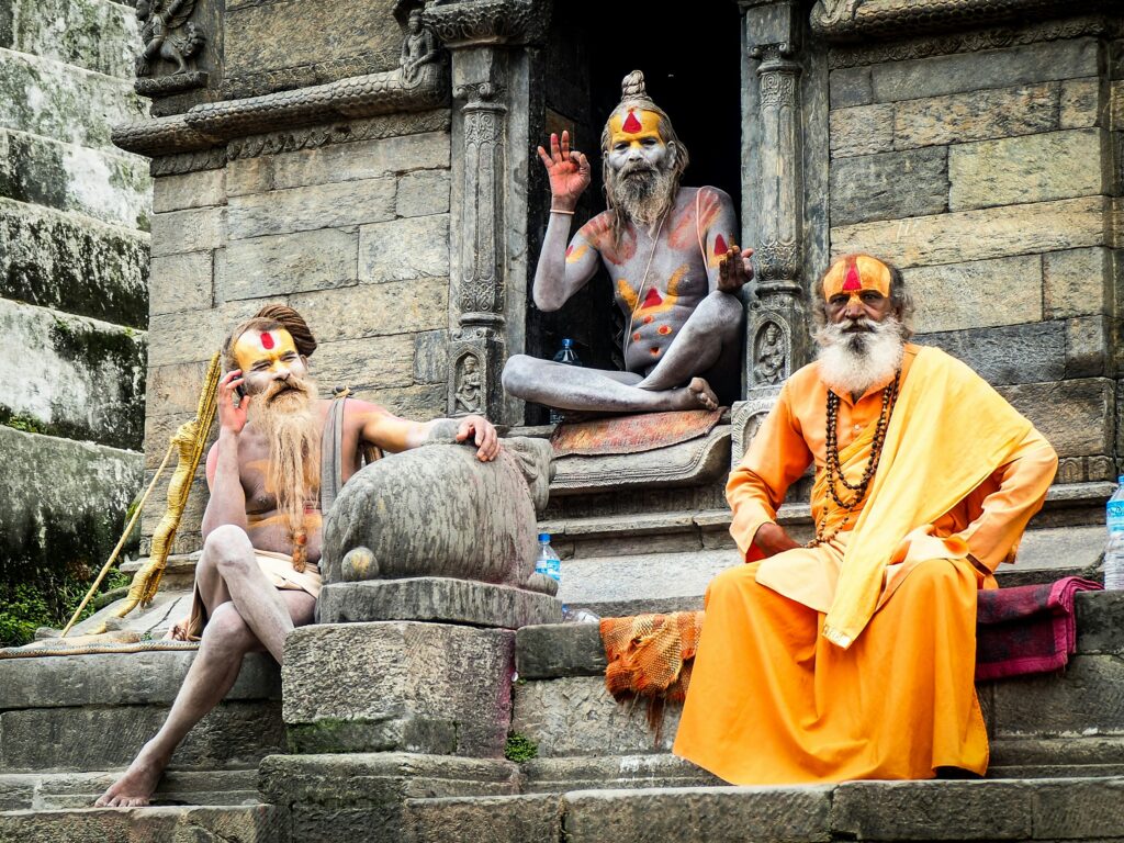 Drei hinduistische Sadhus mit bemalten Gesichtern sitzen auf den Steinstufen eines Tempels. Zwei von ihnen sind nur spärlich bekleidet, einer trägt orangefarbene Gewänder. Im Hintergrund ist ein Steinrelief zu sehen.