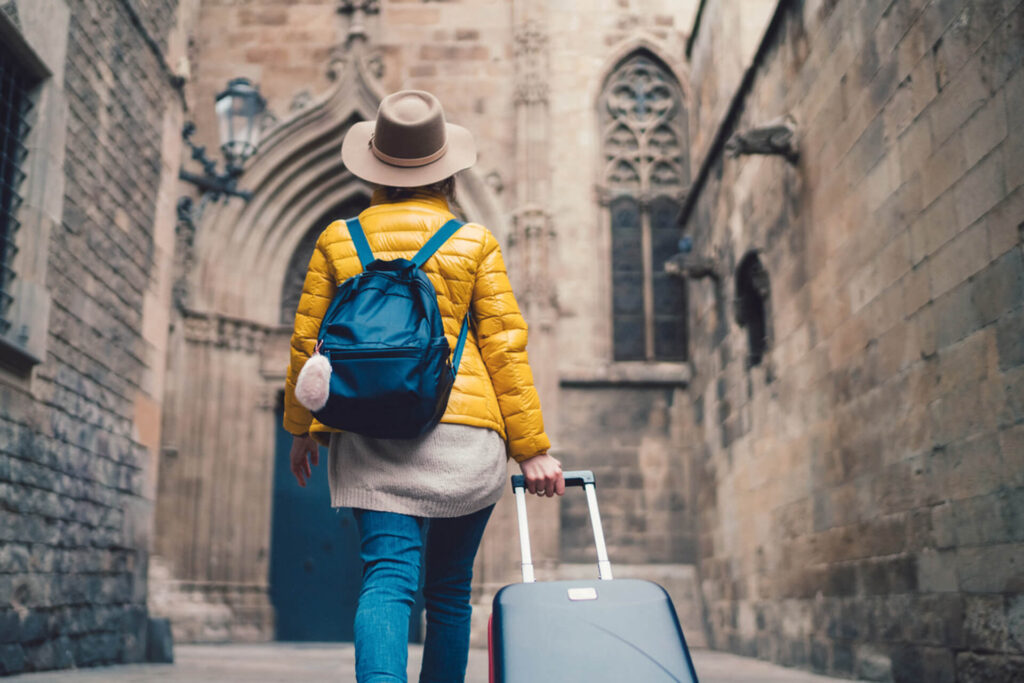 Eine Person in einer gelben Jacke und mit einem Hut, die einen blauen Rucksack und einen Koffer bei sich trägt, geht durch eine historische Gasse aus Stein auf einen Torbogen zu.