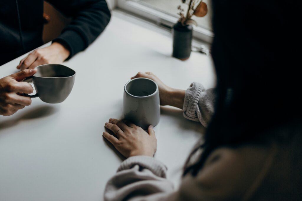 Zwei Personen sitzen an einem Tisch, jede hält eine Tasse in der Hand; im Hintergrund steht eine kleine Topfpflanze auf der Fensterbank.