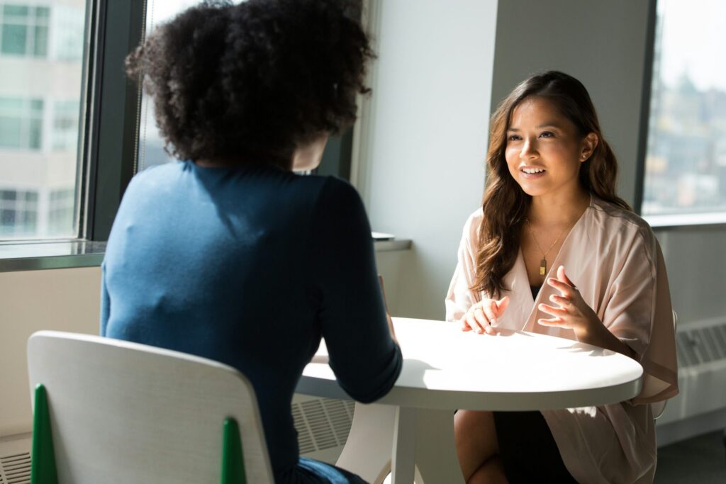 Zwei Frauen sitzen sich an einem runden Tisch am Fenster gegenüber und unterhalten sich in einem hellen, modernen Büro.