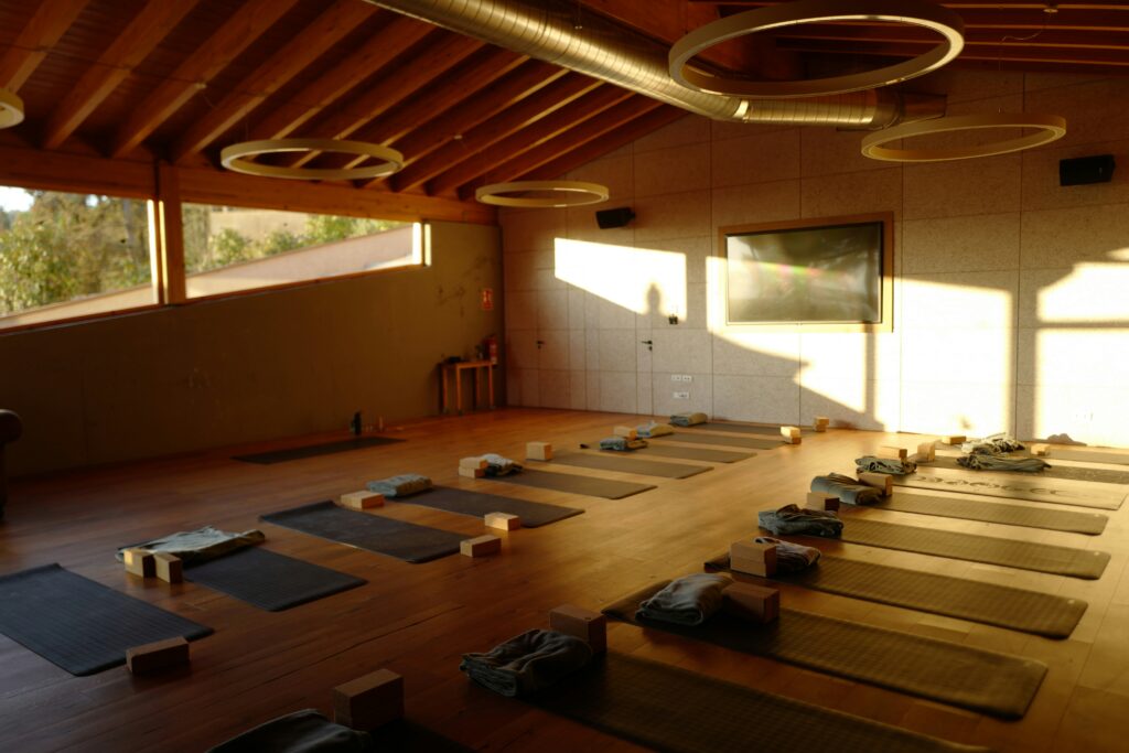 Ein sonnendurchflutetes Yogastudio mit Holzböden, bereits ausgelegten Yogamatten, Blöcken und Handtüchern, runden Deckenleuchten und großen Fenstern, durch die Tageslicht hereinströmt.
