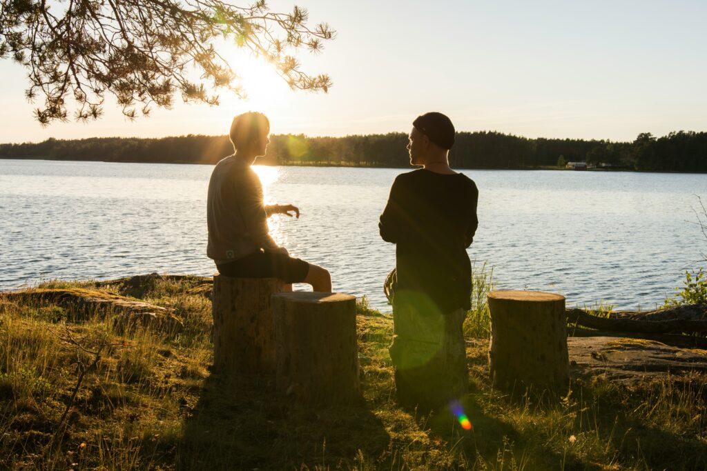 Zwei Menschen sitzen bei Sonnenuntergang auf Baumstümpfen am See und unterhalten sich. Das Sonnenlicht fällt durch die Bäume, zeichnet Silhouetten nach und wirft Schatten auf das Gras.