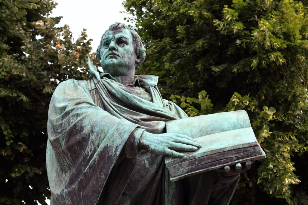 Eine Bronzestatue eines Mannes in einer Robe, der ein aufgeschlagenes Buch in der Hand hält, im Freien aufgestellt, mit grünen Bäumen im Hintergrund.
