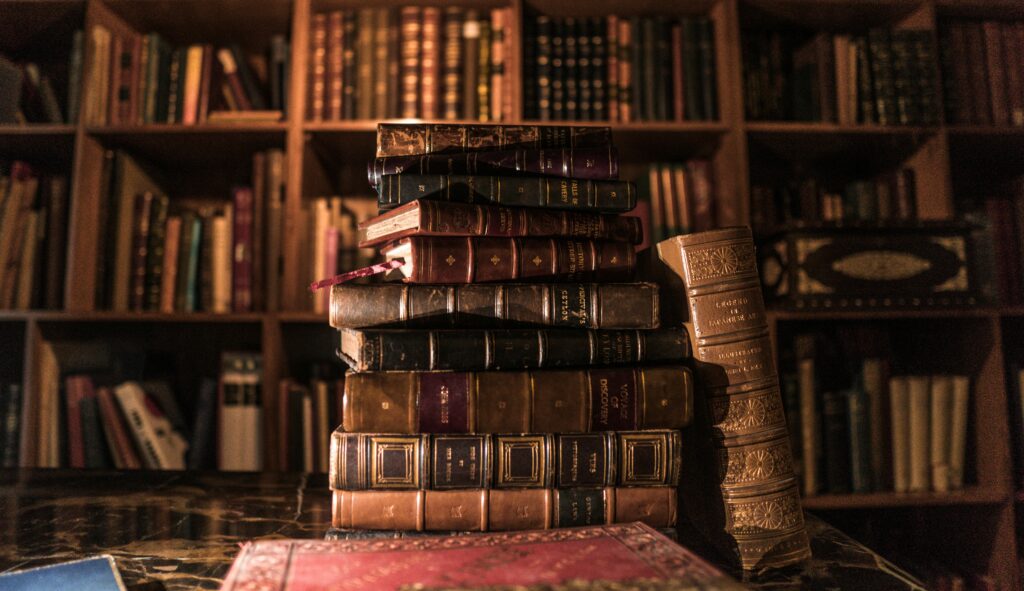 In einer warm beleuchteten Bibliothek steht ein Stapel alter, in Leder gebundener Bücher auf einem Tisch vor einem hölzernen Bücherregal, das mit weiteren Büchern gefüllt ist.