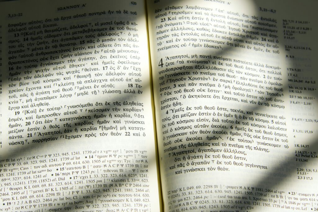 Ein aufgeschlagenes Buch mit griechischem Text wird vom Sonnenlicht beleuchtet, wodurch die Fenstergitter Schatten auf die Seiten werfen. Am unteren Rand sind Fußnoten und Verweisnummern zu sehen.