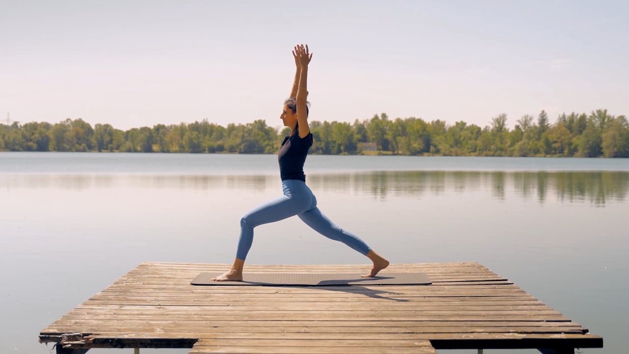 Eine Person praktiziert Yoga in der Krieger-I-Pose auf einem Holzsteg an einem ruhigen See, umgeben von Bäumen unter einem klaren Himmel.