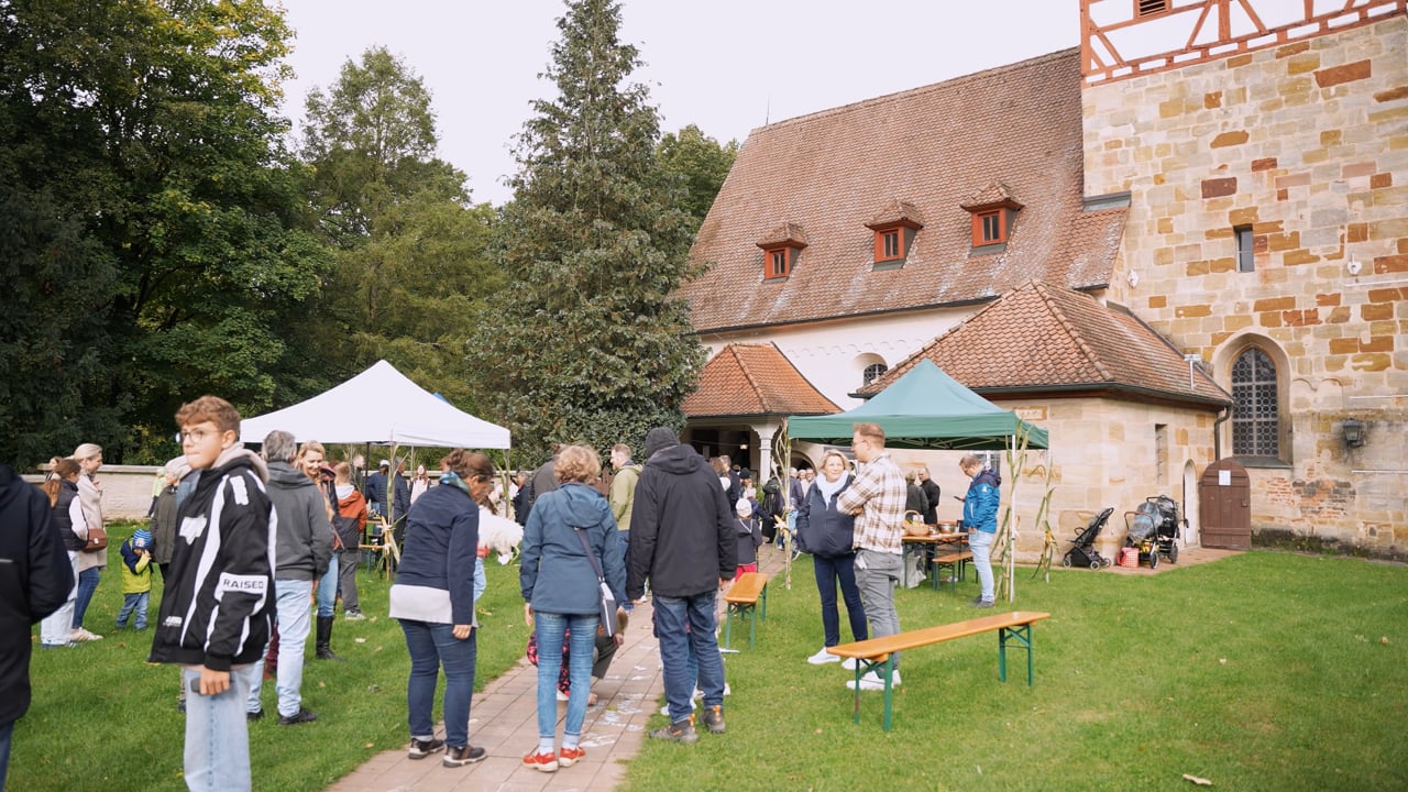 Die Menschen versammeln sich im Freien in der Nähe von Zelten und einem historischen Gebäude zu einer Veranstaltung. Einige stehen im Gespräch, während andere an den Tischen vorbeischauen. Bäume und Gras umgeben die Szene.