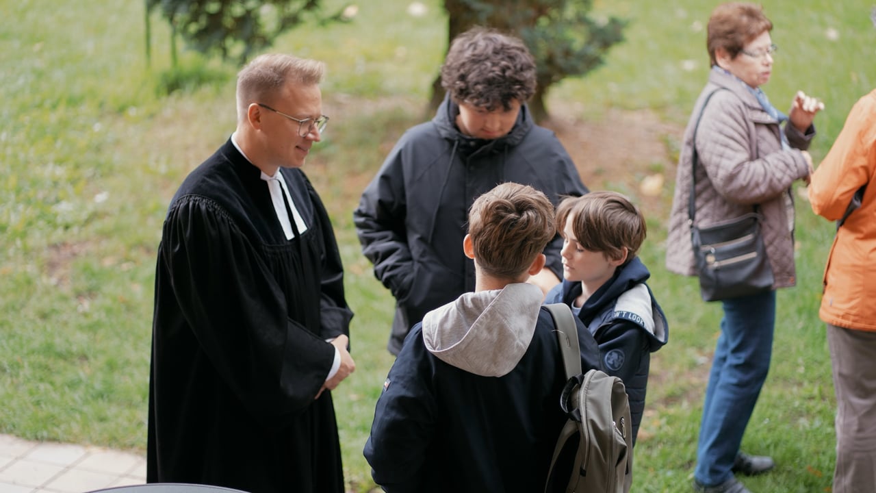 Ein Mann in einer schwarzen Robe unterhält sich im Freien mit drei Jungen, während zwei Frauen in der Nähe stehen. Die Gruppe befindet sich auf einer Rasenfläche, auf der Bäume und ein gepflasterter Weg zu sehen sind.