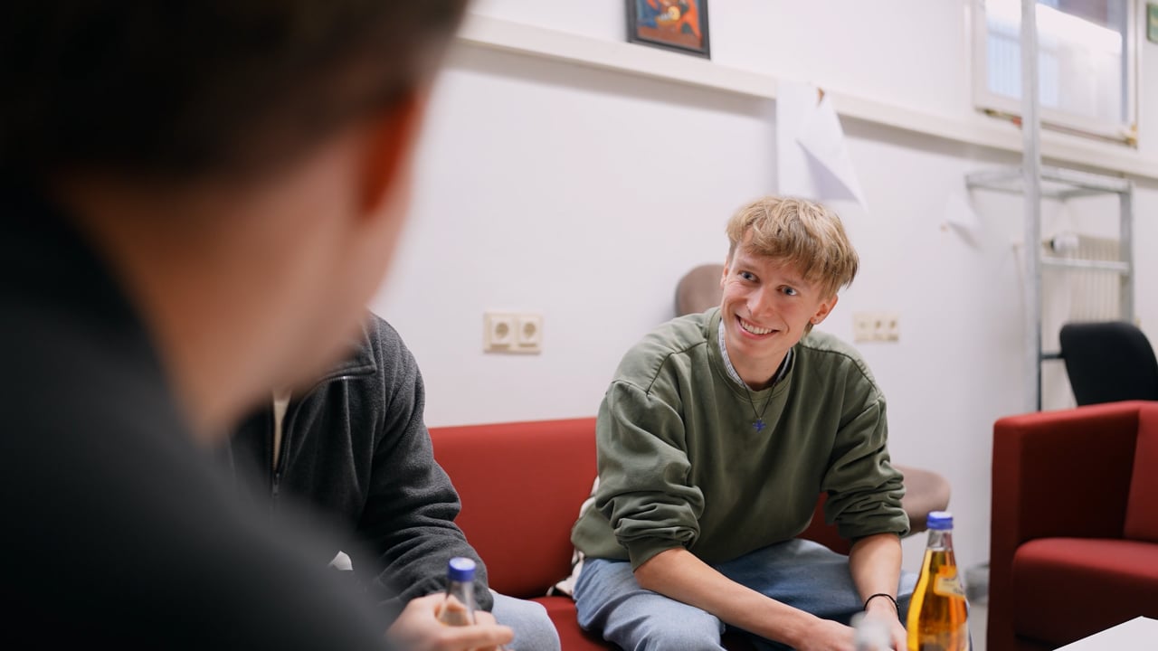 Zwei junge Menschen sitzen auf roten Stühlen und unterhalten sich; einer von ihnen lächelt und hält eine Getränkeflasche in der Hand. Der Fokus liegt auf der lächelnden Person, der Vordergrund ist unscharf und die Szene spielt in einer ungezwungenen Innenraumkulisse.