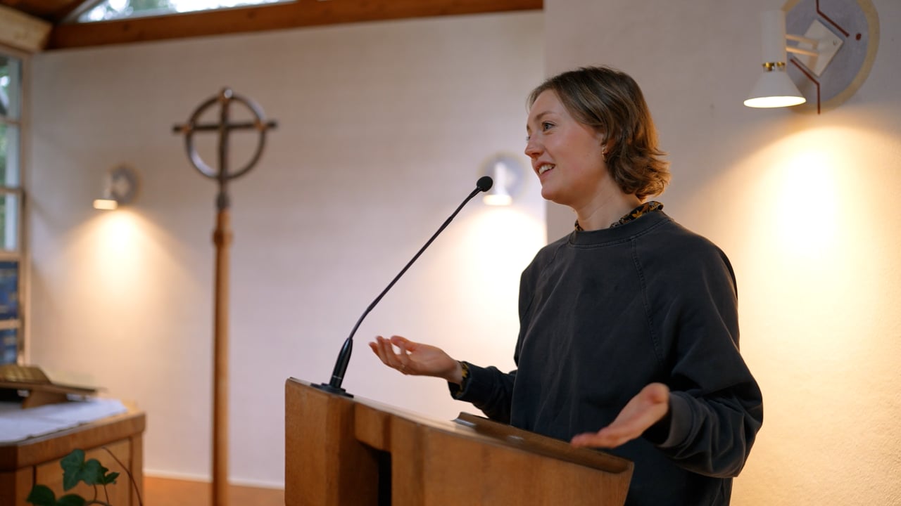 Eine Frau steht an einem Rednerpult und spricht in ein Mikrofon. Sie befindet sich in einem gut beleuchteten Raum, in dessen Hintergrund ein Kreuz und ein Altar zu sehen sind.
