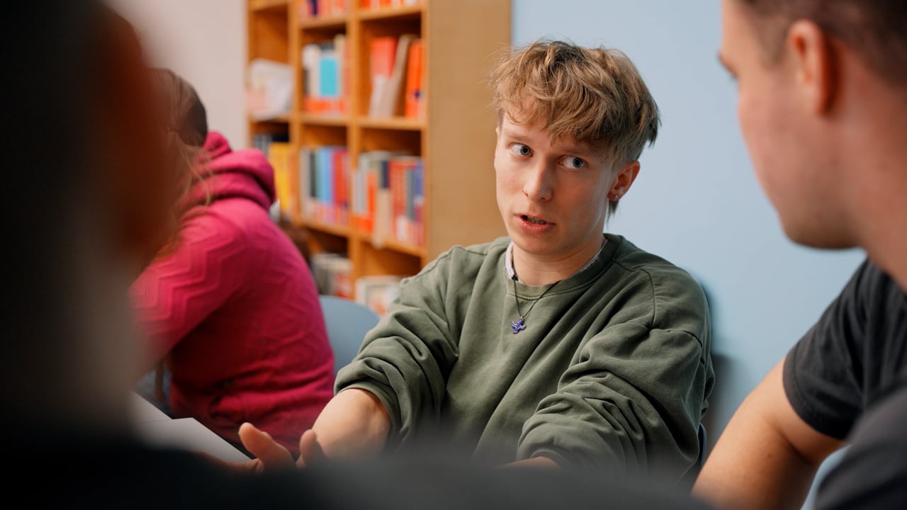 Eine Person mit kurzen Haaren in einem grünen Sweatshirt gestikuliert, während sie sich mit einer anderen Person in einem Klassenzimmer unterhält, im Hintergrund sind Bücherregale zu sehen.