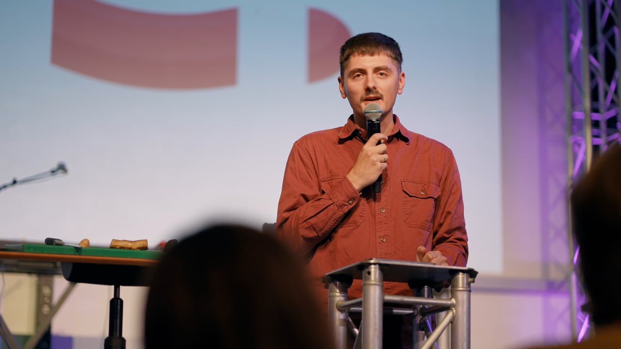 Ein Mann in einem roten Hemd spricht auf der Bühne in ein Mikrofon; er steht hinter einem Rednerpult, im Vordergrund ist ein verschwommenes Publikum zu sehen.