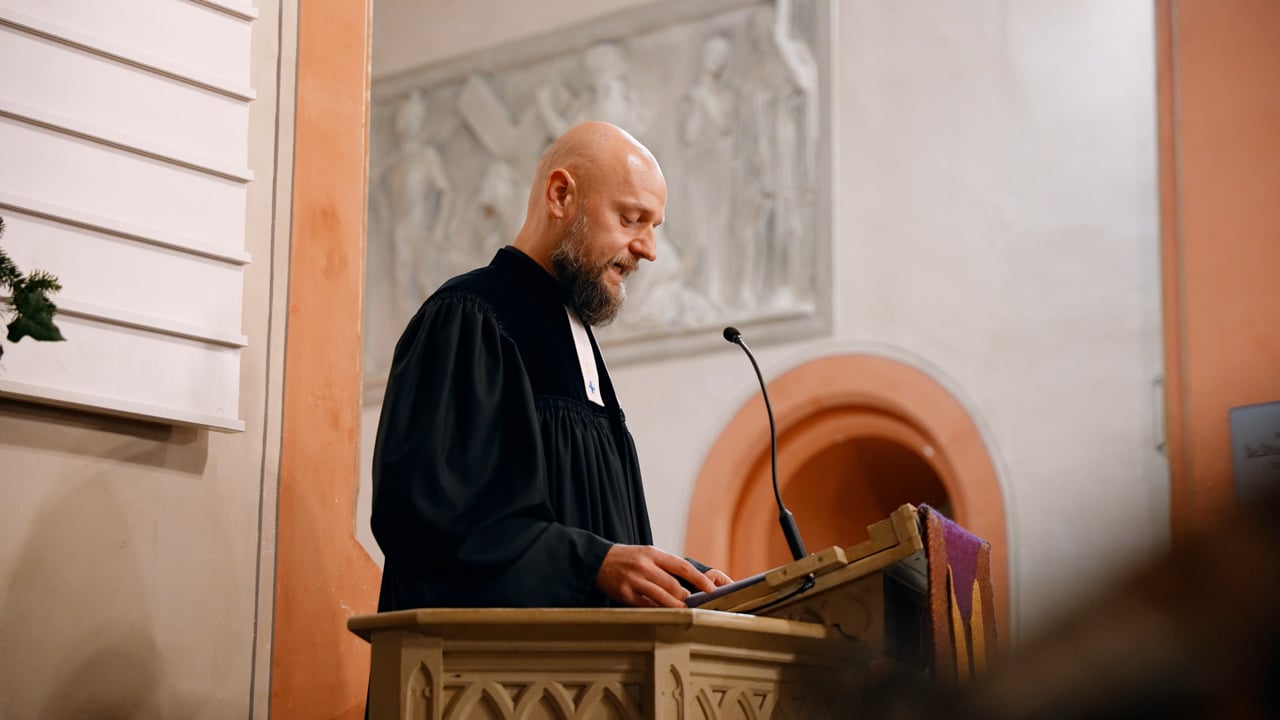 Ein Mann in einer schwarzen Robe steht an einem Rednerpult und spricht in ein Mikrofon in einer Kirche mit pfirsichfarbenen und weißen Wänden.
